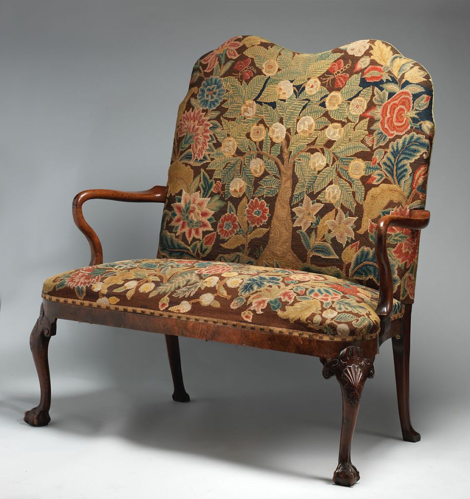 A settee with thin, curved arms and cabriole legs, with the back and seat embroidered with large flowers and leaves surrounding a central tree with white flowers or fruit.